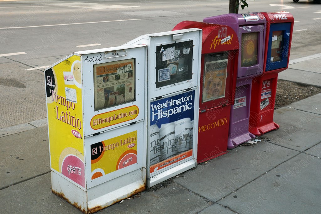 Des distributeurs de journaux hispanophones colorés alignés sur un trottoir.