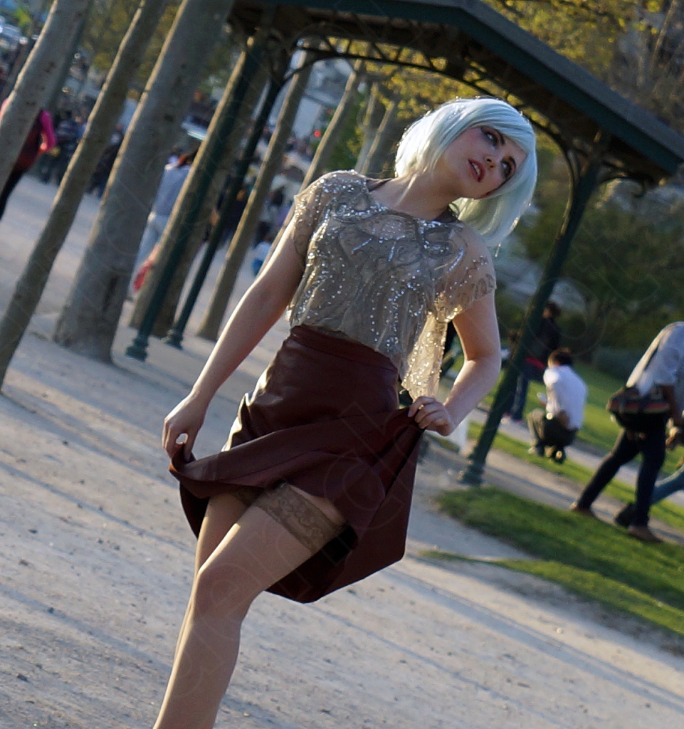 Une jeune femme aux cheveux blond platine, en jupe bordeaux, pose dans une allée du Champ-de-Mars.