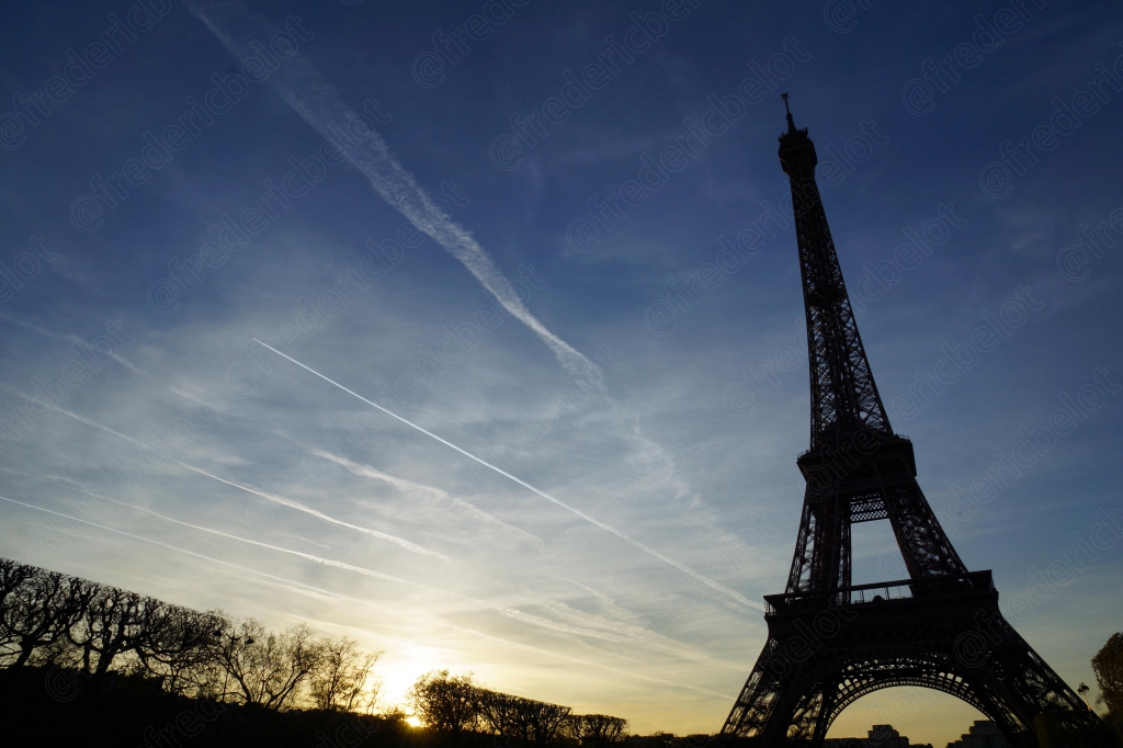 La tour Eiffel se découpe à contre-jour dans un ciel strié de traînées de condensation, au lever du jour.
