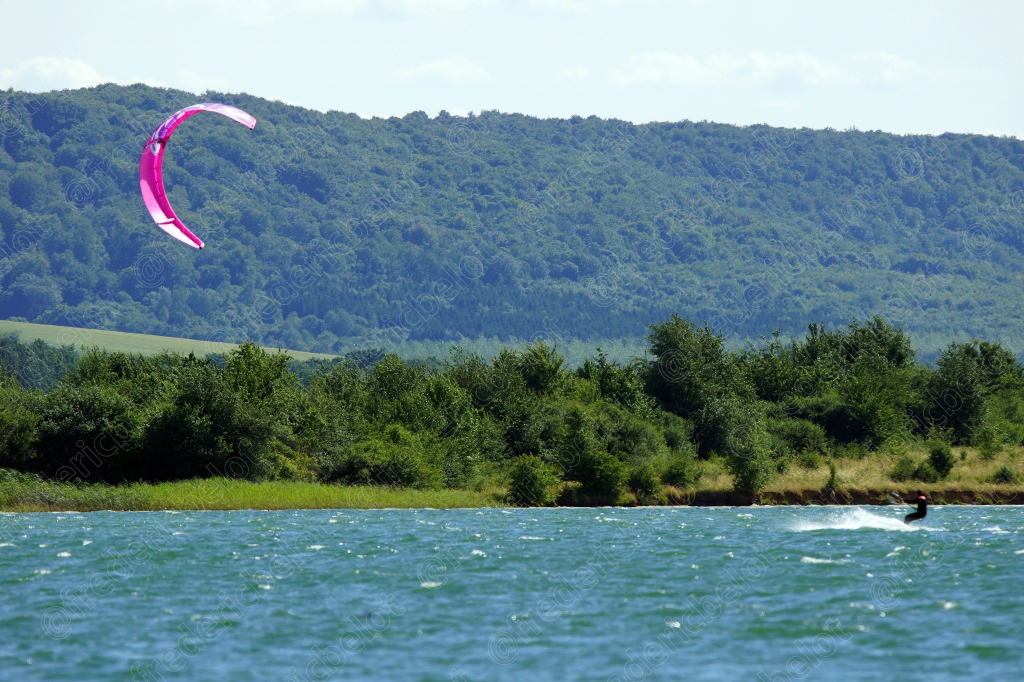 Un kitesurfeur glisse sur un lac bordé de collines boisées, aile rose déployée dans le ciel.