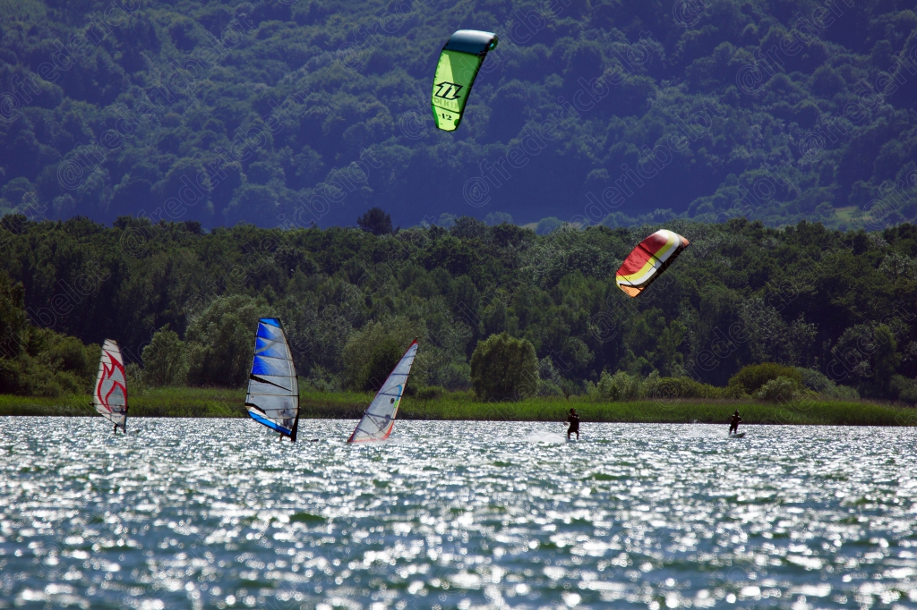 Plusieurs kitesurfeurs et véliplanchistes évoluent sur un plan d'eau scintillant de soleil.
