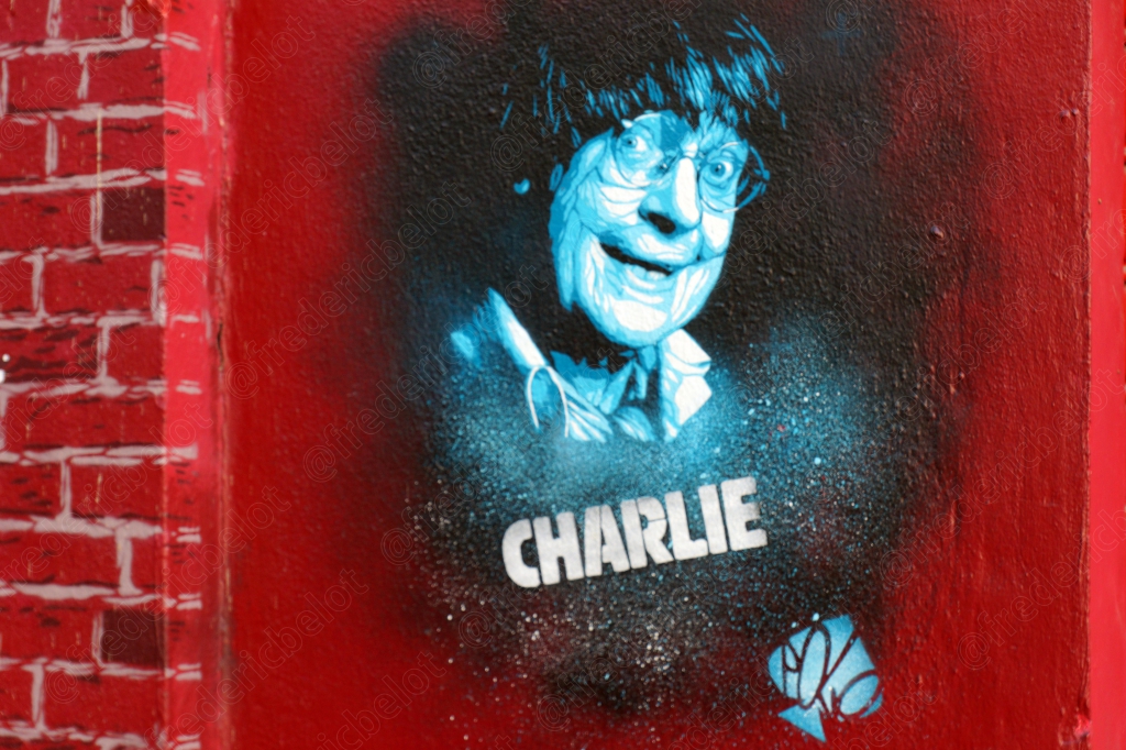 Un pochoir de street art bleu et blanc représentant un visage stylisé, accompagné du mot "Charlie", sur un mur de brique rouge.