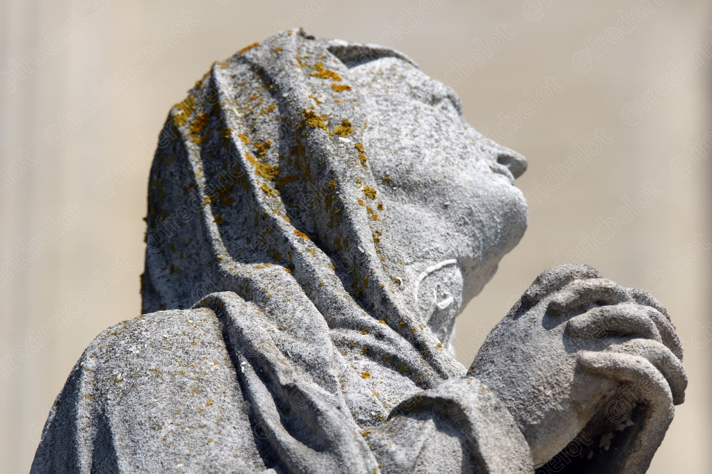 Une statue de pierre représentant un visage levé vers le ciel, mains jointes en prière.