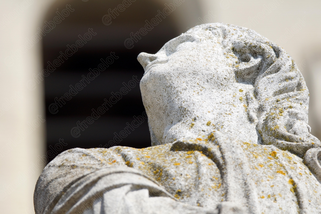 Une statue de pierre vue de dos, capuche relevée, dans une posture recueillie.