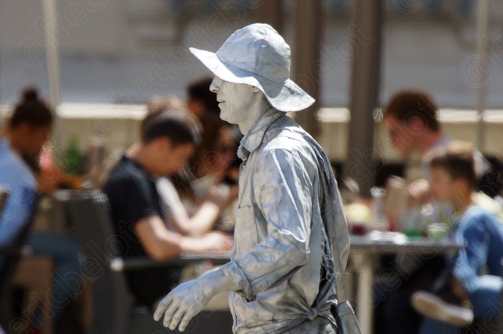 Une statue humaine costumée en argent se tient immobile parmi les terrasses de café animées.