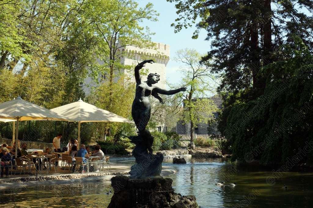 Une fontaine surmontée d'une sculpture en bronze, dans un jardin ombragé bordé de terrasses.