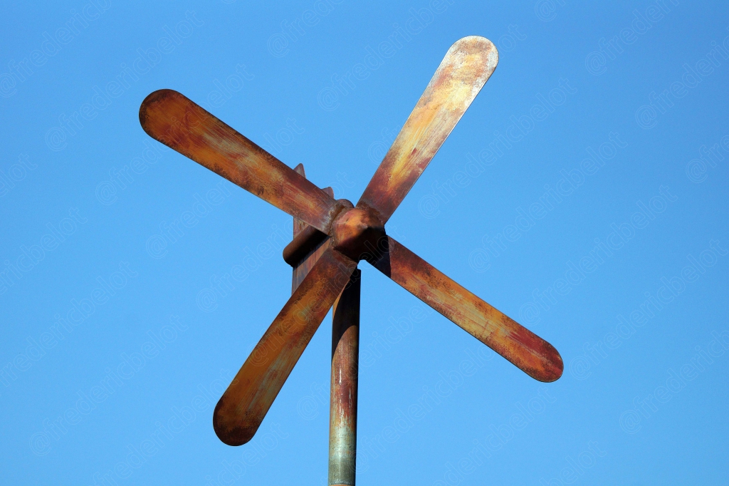 Une girouette artisanale en bois se découpe sur un ciel bleu uni.