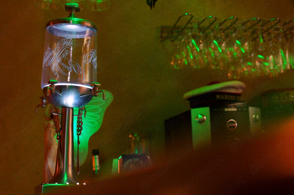 Ambiance colorée d'un comptoir de bar la nuit, bouteilles et lumières vertes et rouges.