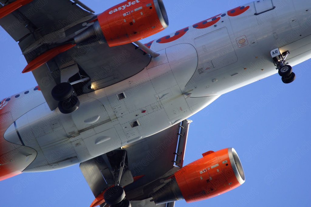 Un avion easyJet vu de dessous au moment de l'atterrissage, dans un ciel bleu.