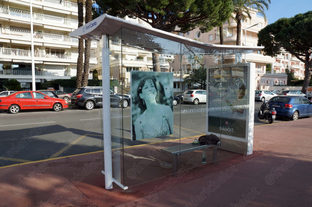 Un arrêt de bus habillé d'une affiche artistique, le long d'une avenue bordée de palmiers.