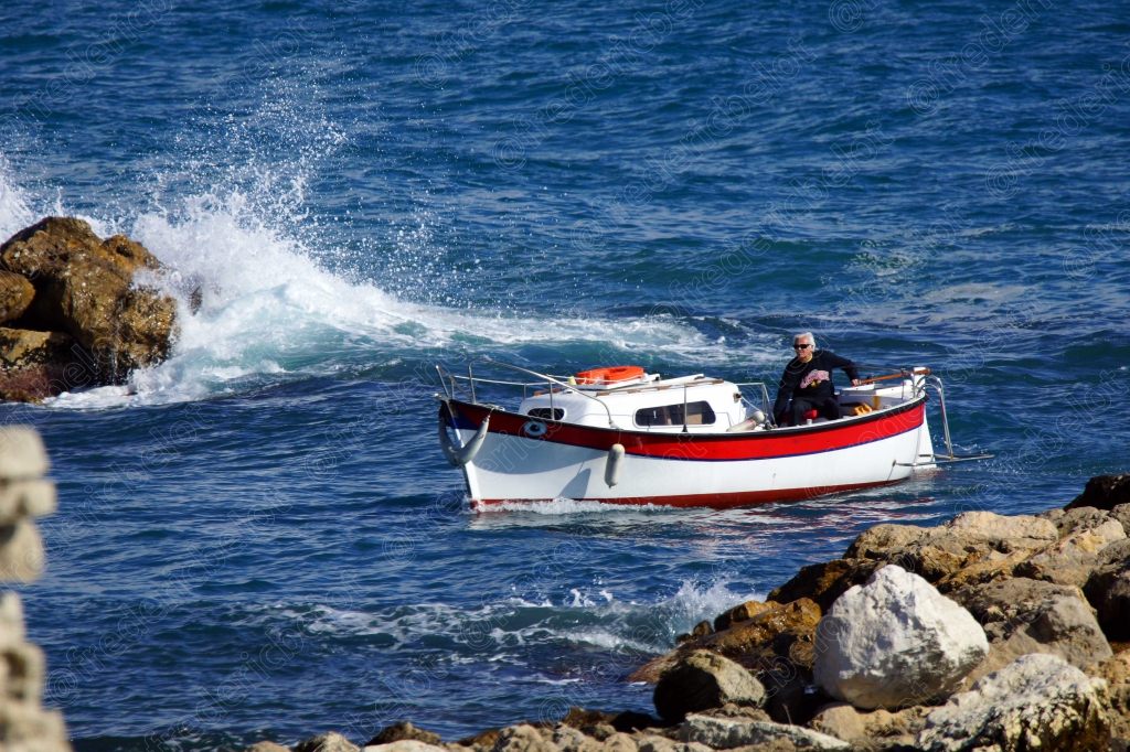 Un petit bateau de pêche navigue près des rochers, dans les embruns d'une vague qui se brise.