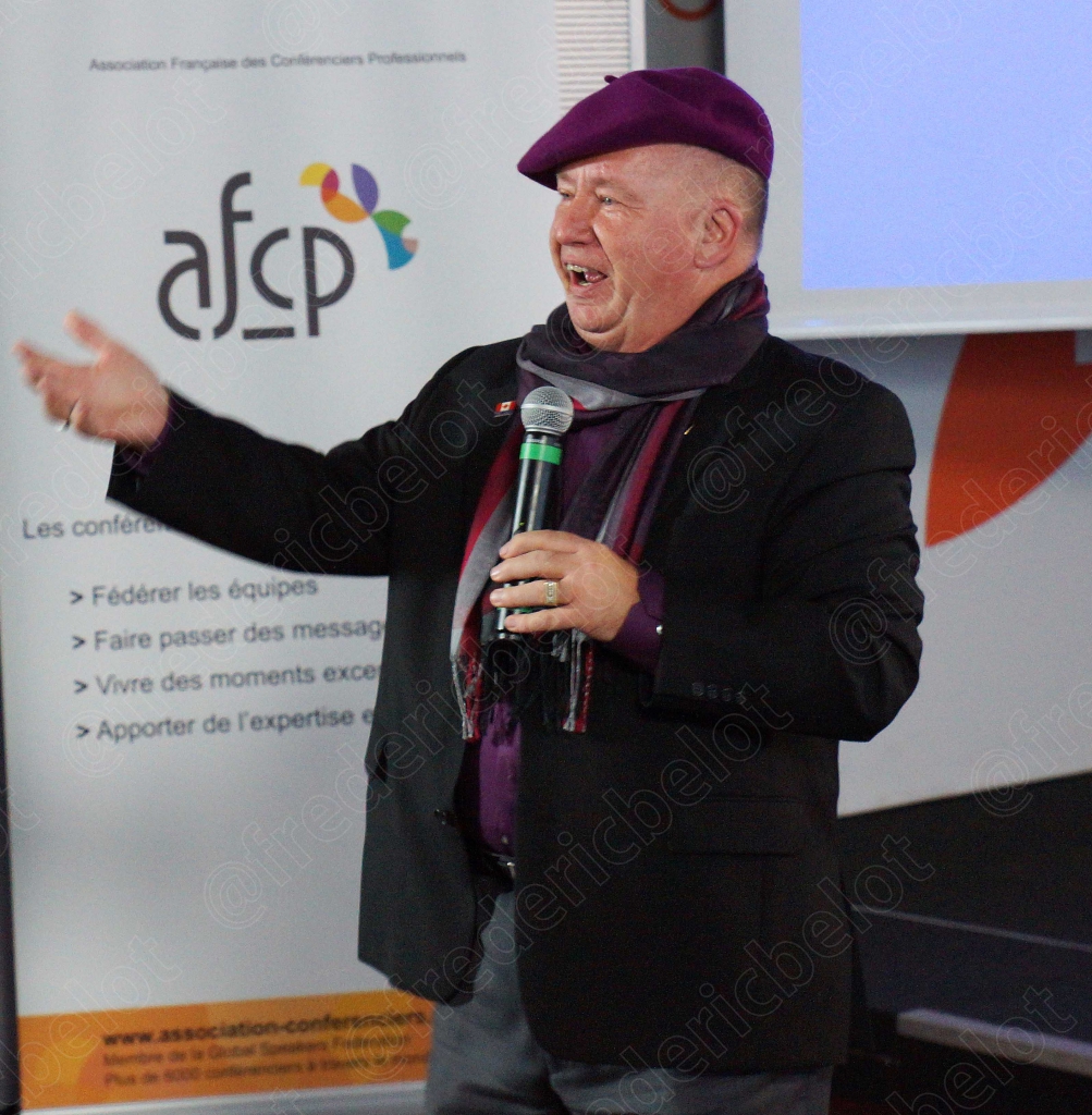 Un homme coiffé d'un béret violet s'exprime au micro, bras tendu, devant le kakémono AFCP.