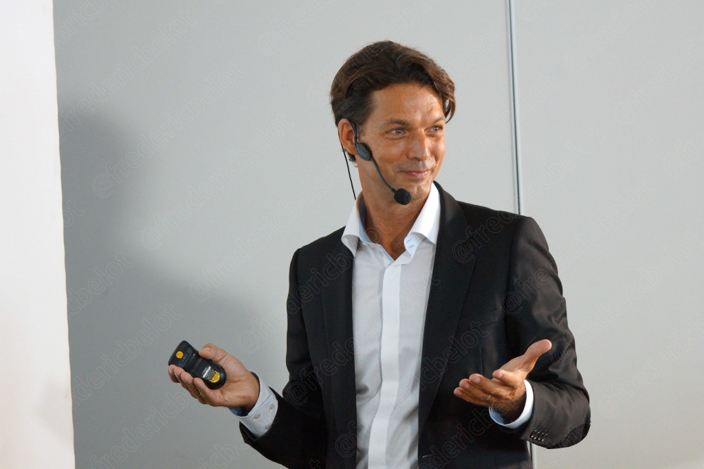 Un homme en costume sombre s'exprime, télécommande de présentation en main.