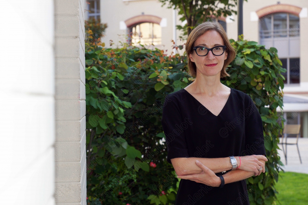 Portrait d'une femme en tenue noire, bras croisés, posant devant un jardin et une façade en pierre.
