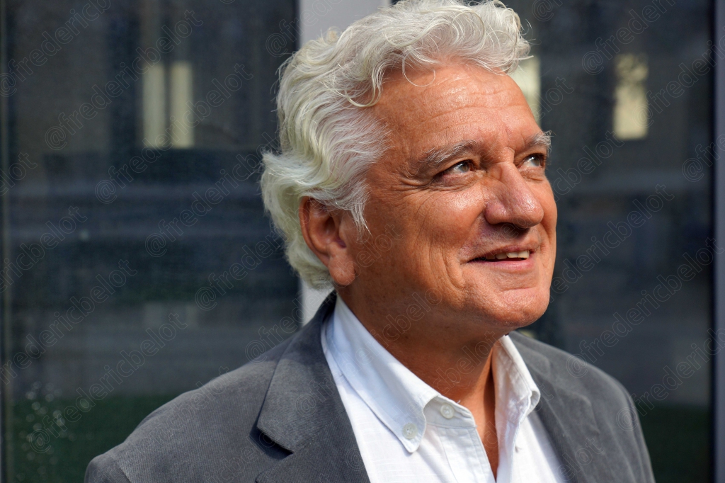 Portrait d'un homme aux cheveux gris et veste claire, souriant de profil trois-quarts.