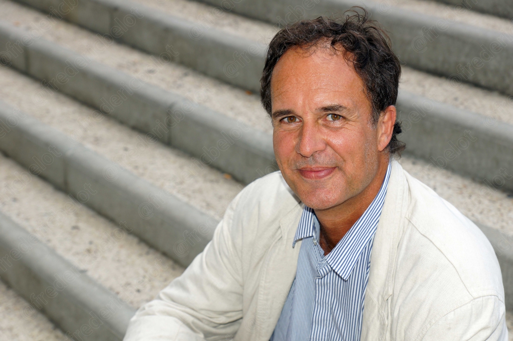 Portrait d'un homme assis sur des marches en pierre, veste claire, souriant à la caméra.