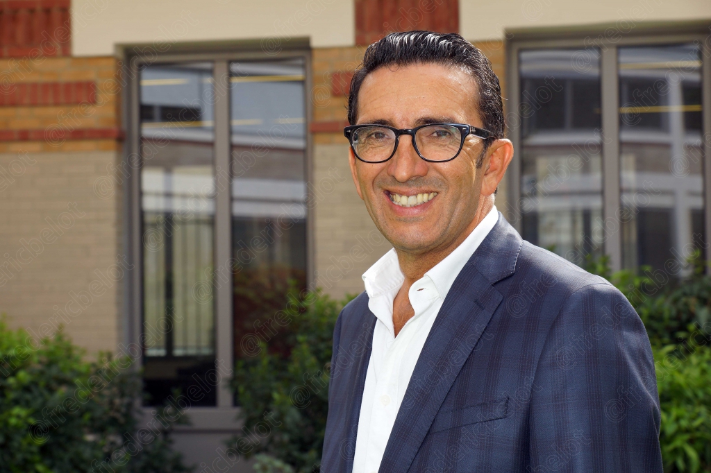 Portrait souriant d'un homme à lunettes, costume sombre et chemise blanche, devant une façade en pierre et brique.