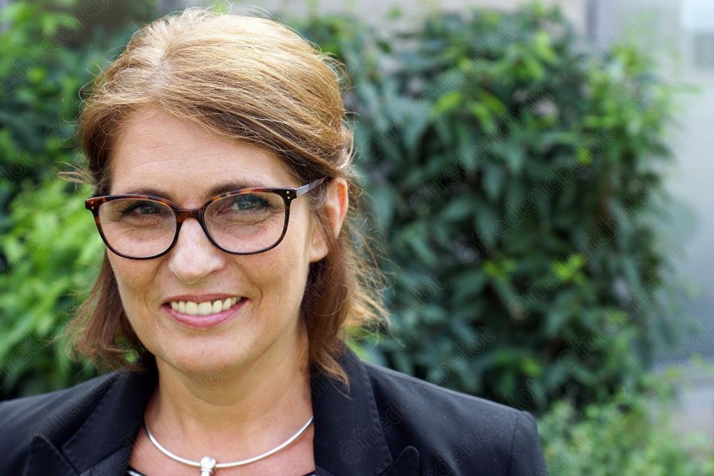 Portrait souriant d'une femme à lunettes, cheveux blond-roux, devant un feuillage vert.
