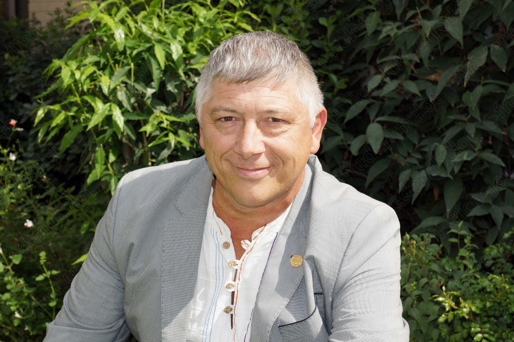 Portrait souriant d'un homme aux cheveux gris, veste claire, devant un feuillage dense.