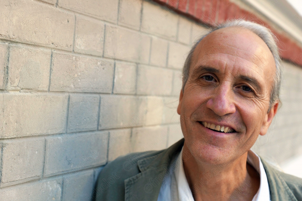 Portrait souriant d'un homme aux cheveux gris, en veste, devant un mur en parpaings clairs.
