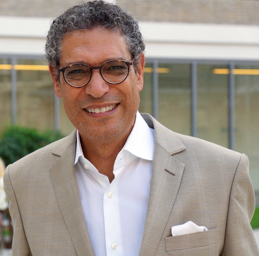 Portrait souriant d'un homme à lunettes, veste beige, devant une façade vitrée.
