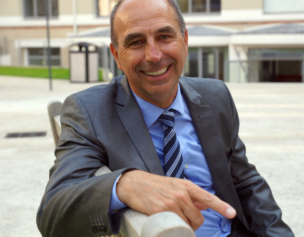 Portrait d'un homme en costume, souriant, assis et pointant du doigt, dans une cour extérieure.
