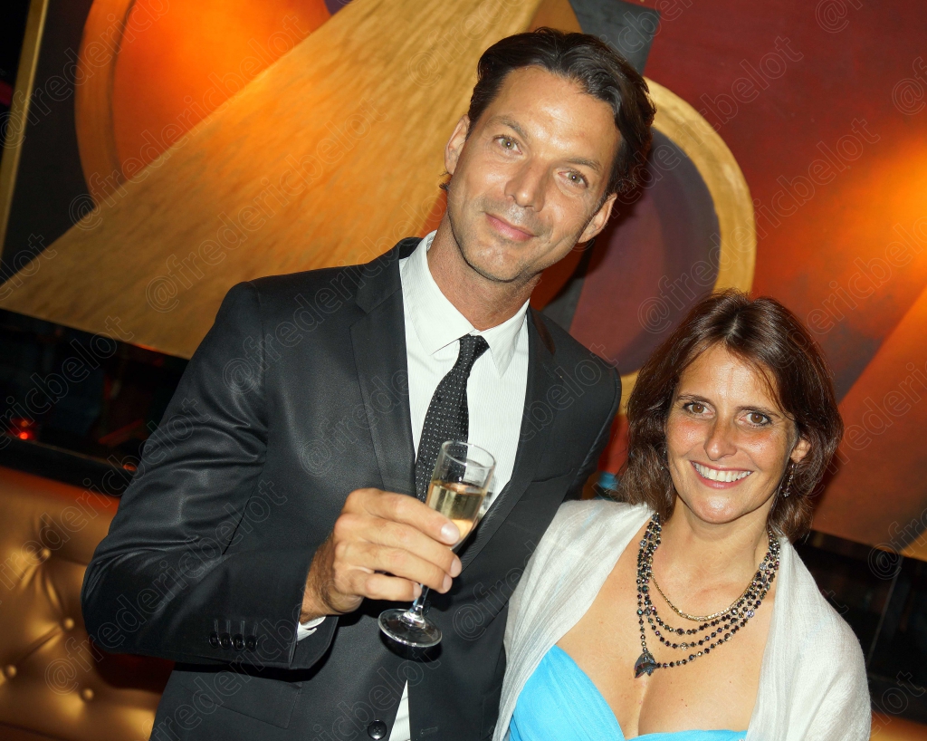 Un homme en costume sombre et une femme en robe bleue et blanche posent, coupe de champagne à la main.