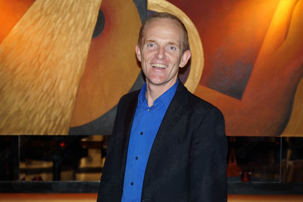 Portrait d'un homme en chemise bleu nuit, souriant devant le photo call.