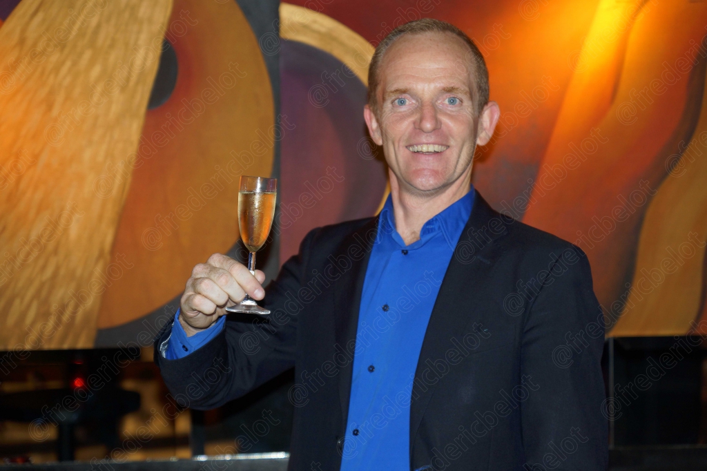 Un homme en chemise bleue lève sa coupe de champagne devant le photo call.
