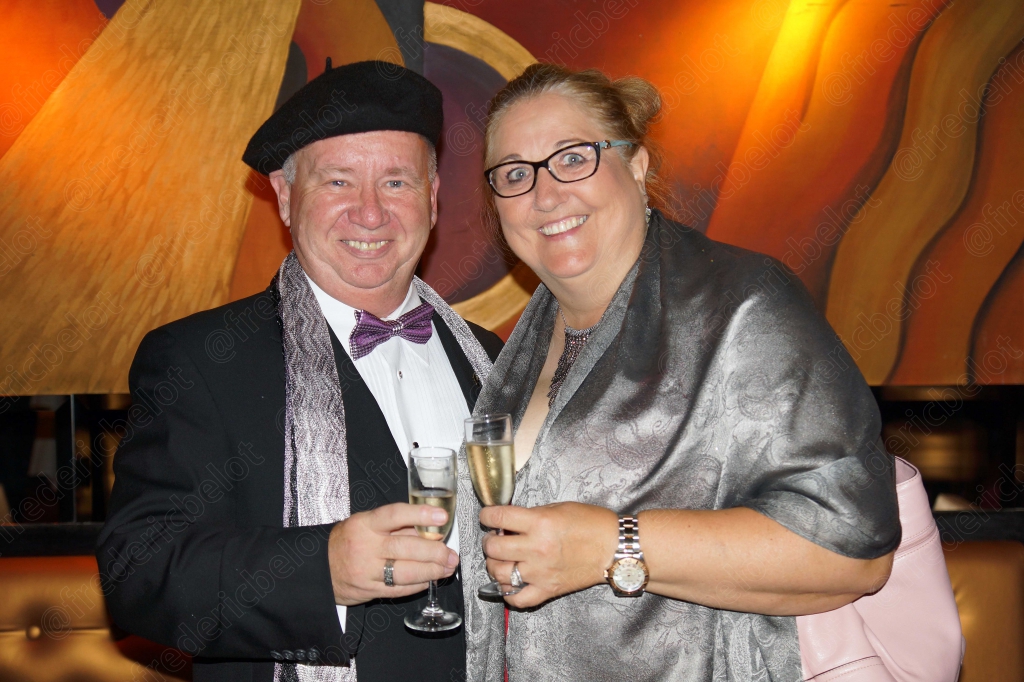 Un homme en béret et une femme à lunettes posent ensemble, coupes de champagne en main.