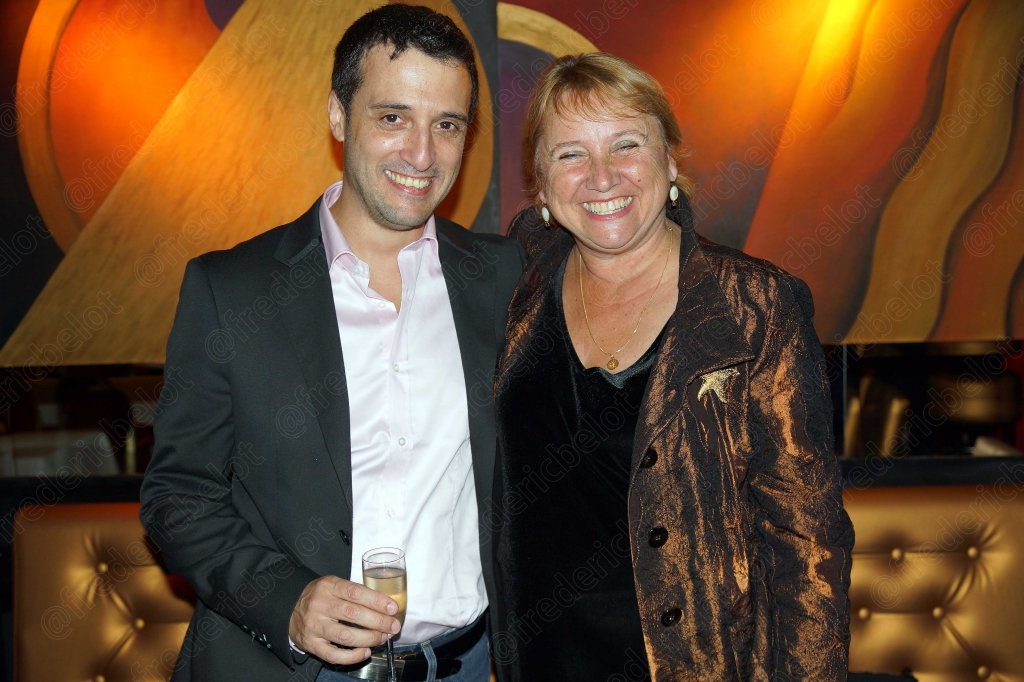 Un homme en chemise blanche et une femme en noir posent, coupe de champagne en main.