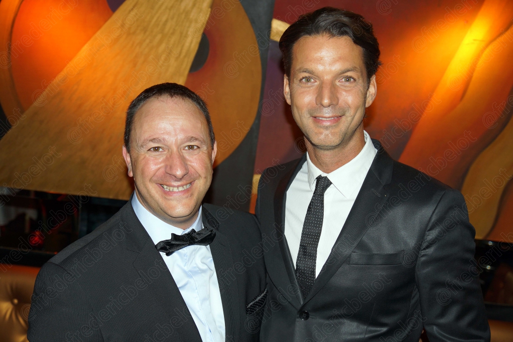 Deux hommes en costume et nœud papillon posent ensemble, souriants.