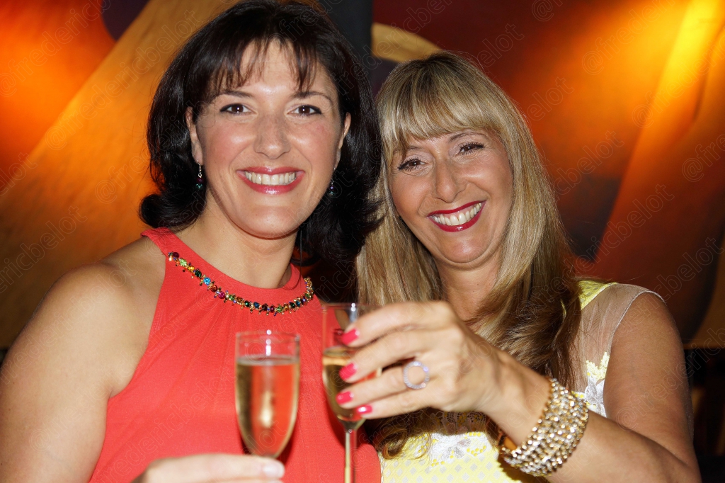 Deux femmes en robe rouge et jaune trinquent ensemble, souriantes.