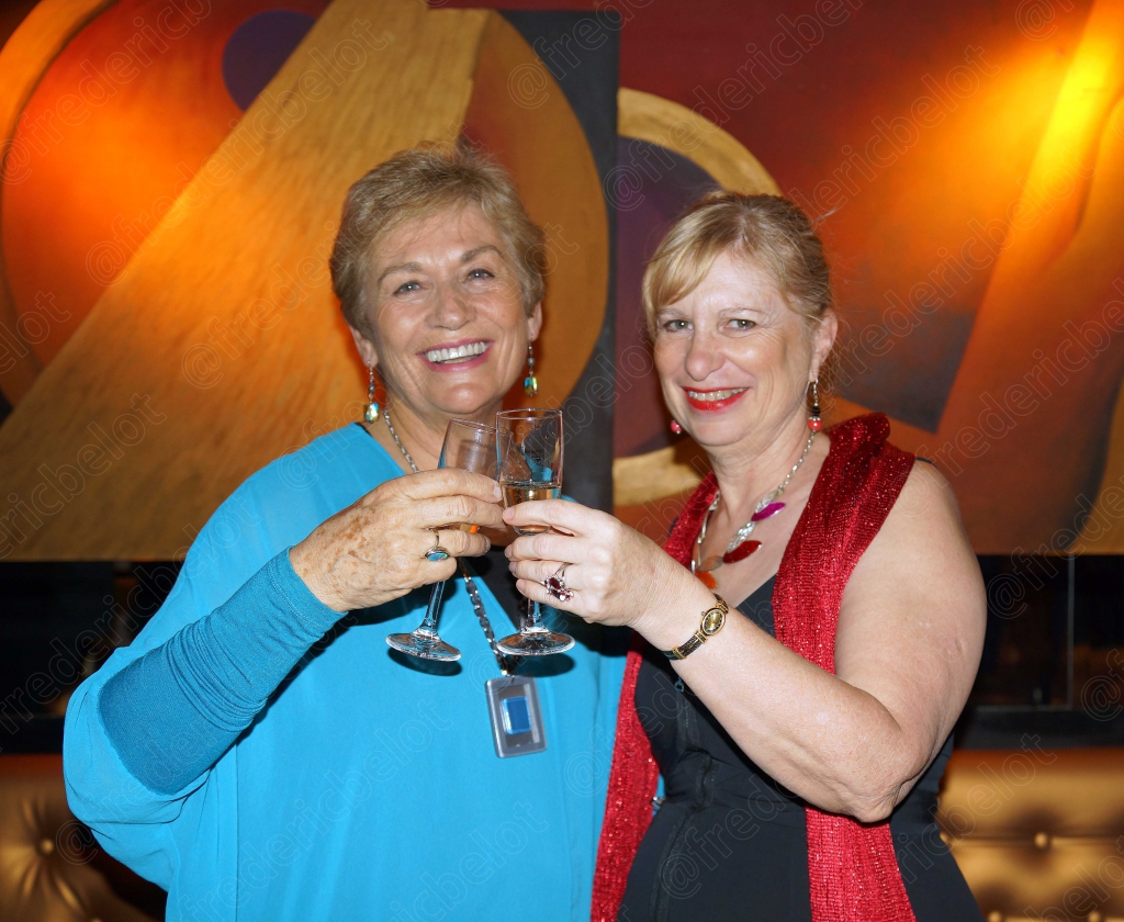 Deux femmes en robe bleue et rouge trinquent ensemble, souriantes.