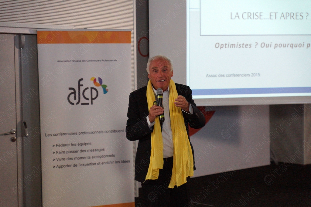 Un homme en costume, foulard jaune autour du cou, parle au micro devant la diapositive "La crise... et après ?".