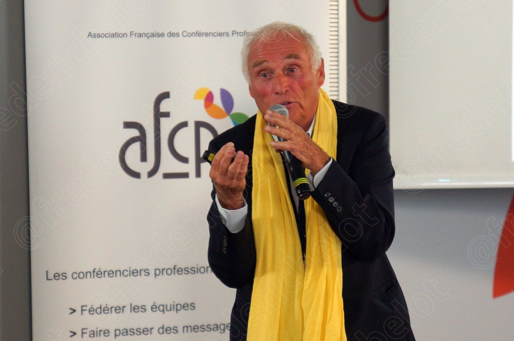 L'intervenant au foulard jaune gesticule, mains ouvertes, devant le kakémono AFCP.