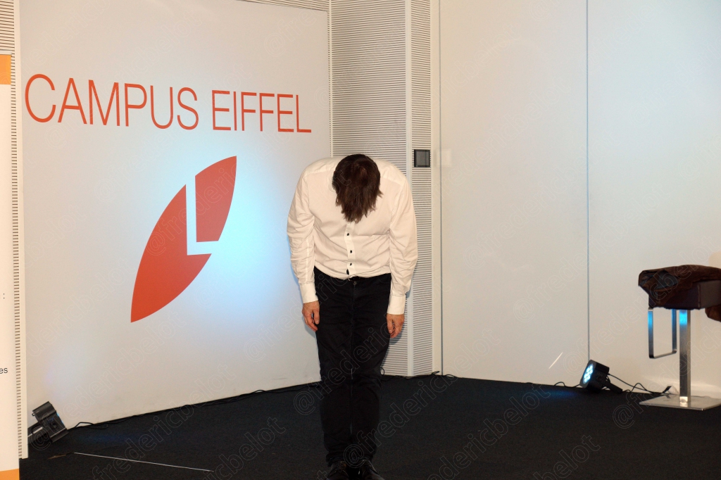 Un homme en chemise rose s'exprime avec animation devant le kakémono AFCP éclairé de bleu.
