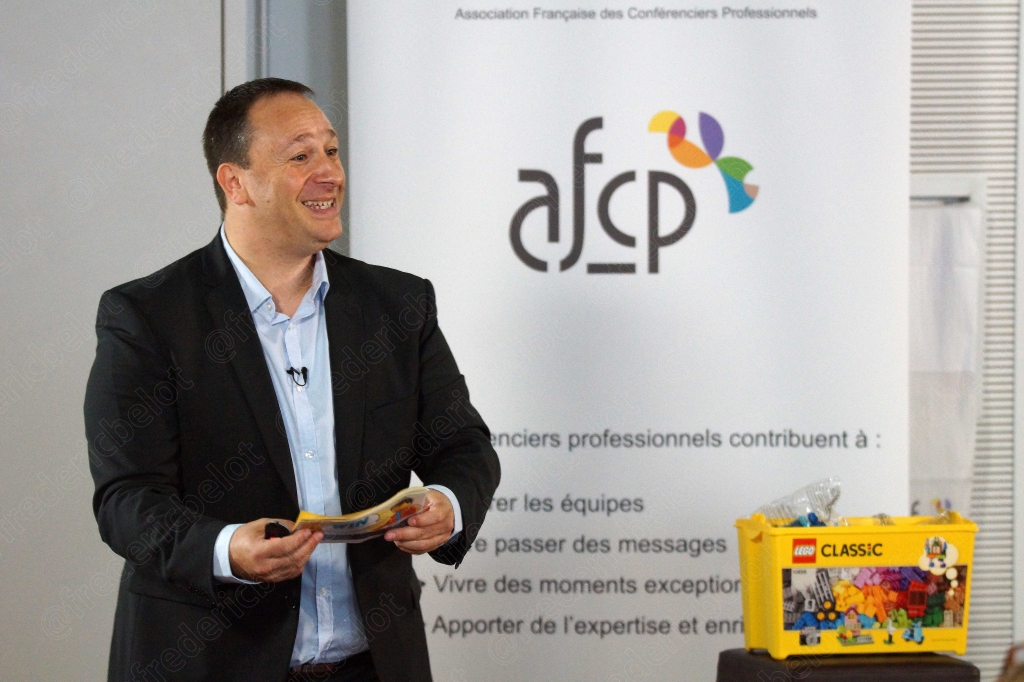 Homme en costume tenant une boîte de jeu de construction, devant un kakémono afcp