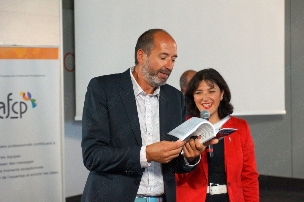 Homme et femme en veste rouge sur scène, elle tient un livre