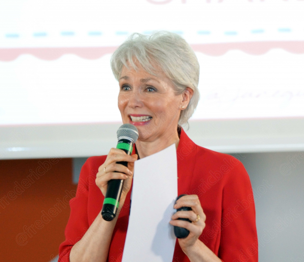 Femme en veste rouge parlant au micro, écran de projection en fond