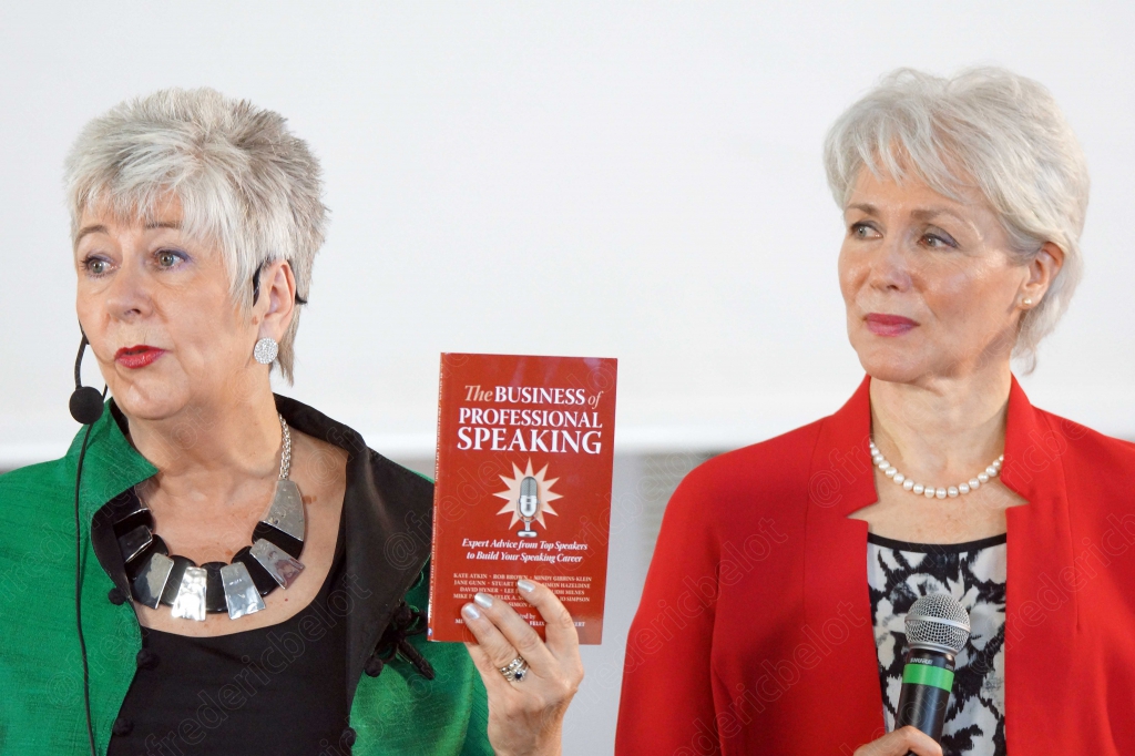 Deux femmes en veste verte et rouge sur scène, l'une tient un livre sur l'art de la conférence