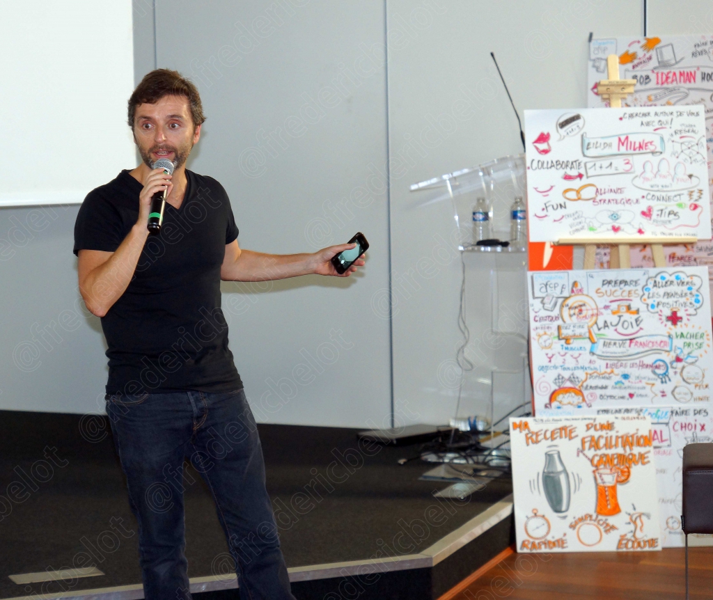 Homme en t-shirt noir parlant au micro, tablette en main, devant un mur de mind-mapping
