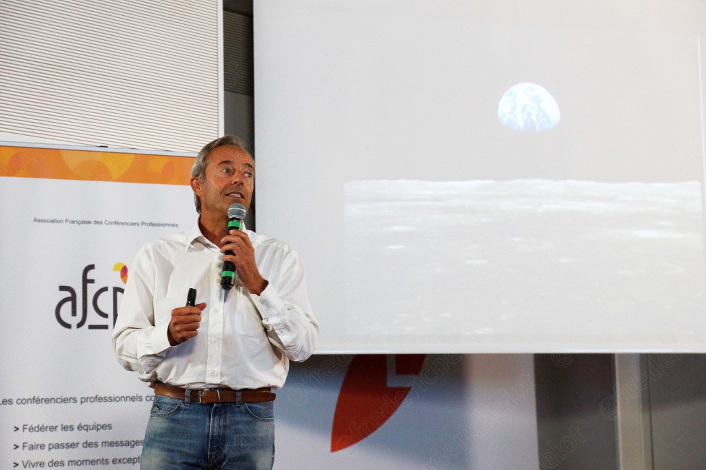 Homme en chemise blanche parlant au micro, écran affichant une image spatiale