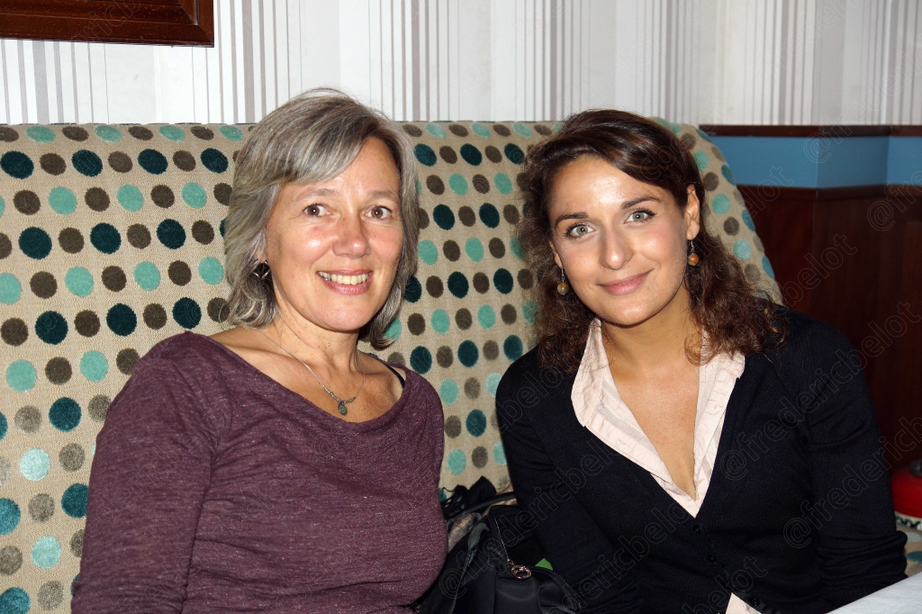 Deux femmes souriantes posent ensemble à table, banquette à motifs en arrière-plan.
