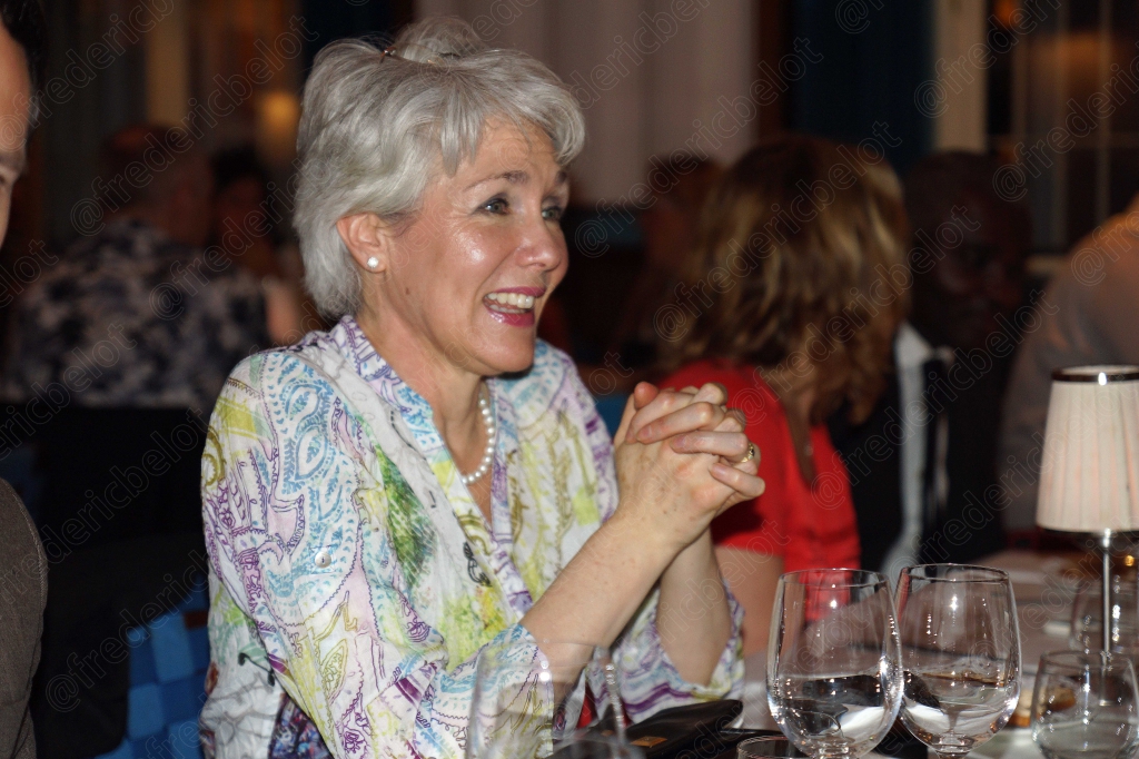 Une femme aux cheveux argentés rit, mains jointes, à table.