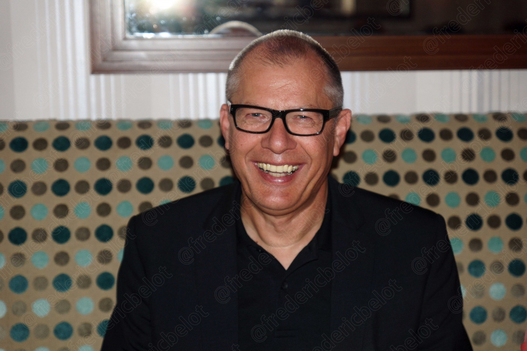 Portrait souriant d'un homme à lunettes, chemise sombre, devant un mur à motifs ronds colorés.