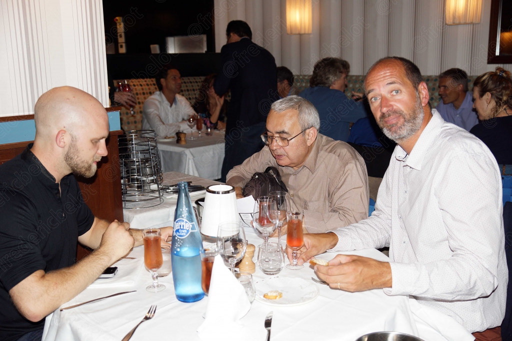 Trois hommes discutent autour d'une table, bouteille d'eau et verres à cocktail devant eux.