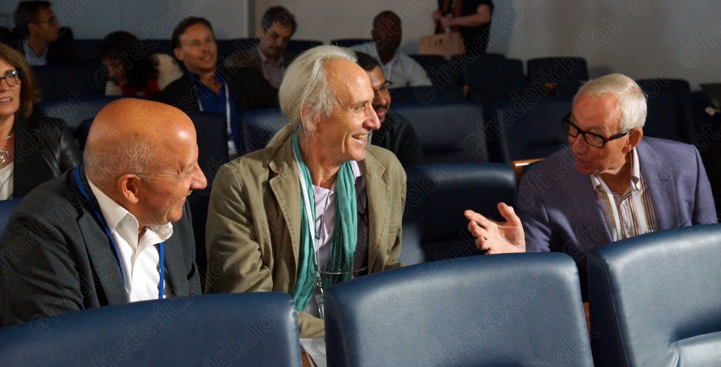 Trois hommes assis dans le public, l'un d'eux souriant largement.
