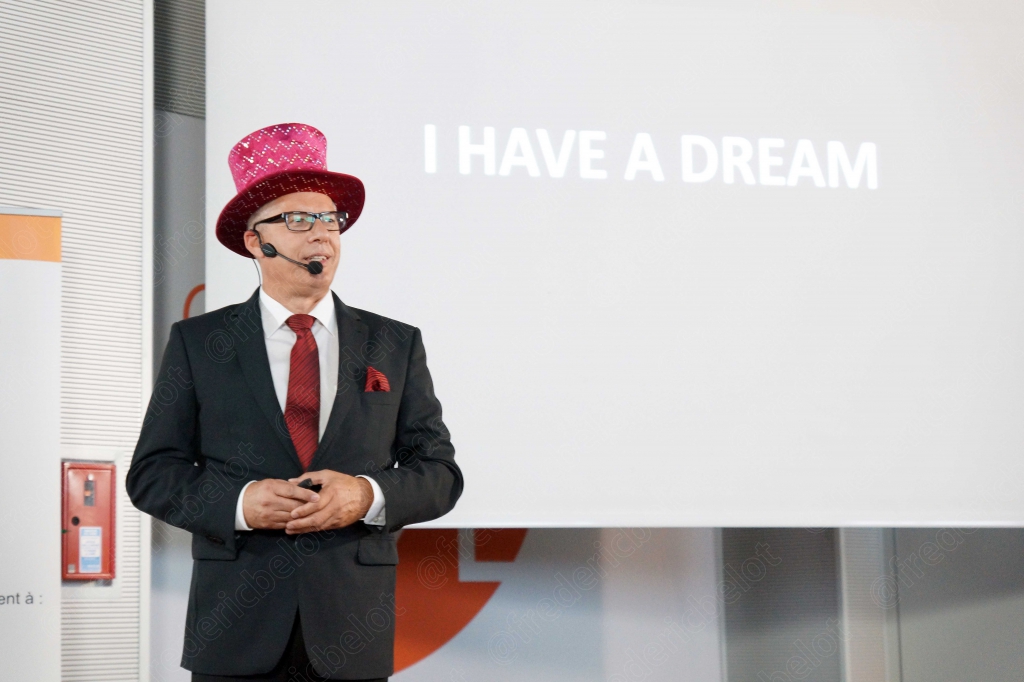 L'intervenant au haut-de-forme rose se tient devant une diapositive "I have a dream".