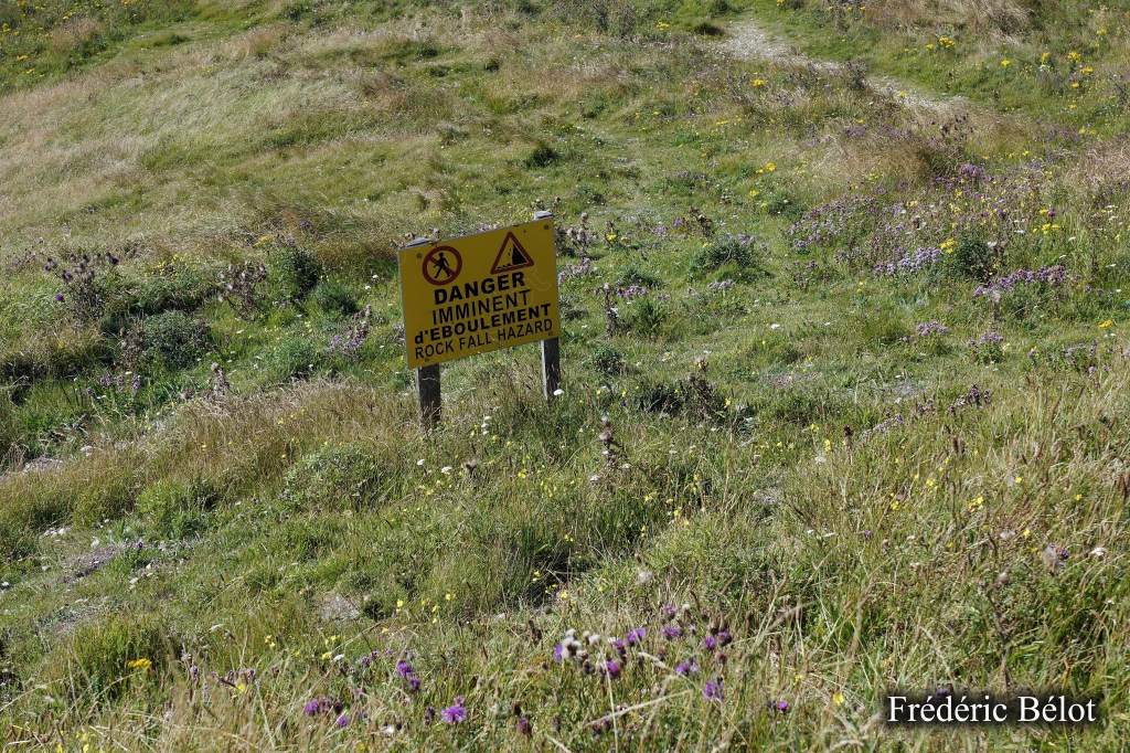 Le soleil perce entre les nuages au-dessus d'une prairie côtière, panneau de danger visible.